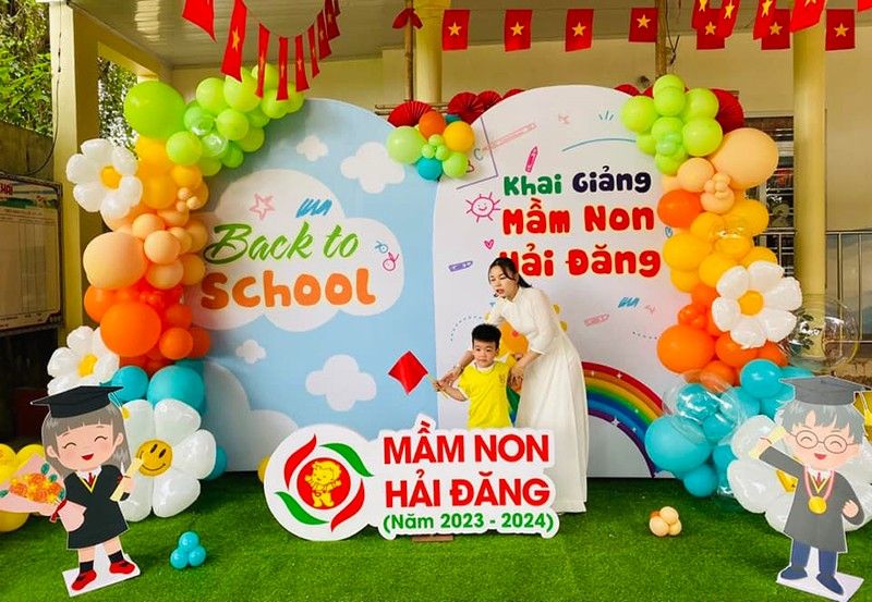 2 người chụp hình bên backdrop khai giảng trường mầm non